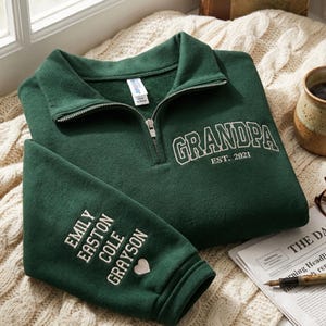 May include: A dark green sweatshirt with a quarter-zip collar. The word "GRANDPA" is embroidered in white, along with "EST. 2021". The left sleeve has embroidered names and a heart symbol. The sweatshirt is folded on a cream-colored knitted blanket.