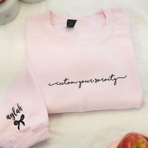 Może przedstawiać: Jasnoróżowa bluza z napisem "custom your sorority" czarnym skryptem. Lewy rękaw ma imię "aylah" wyhaftowane czarnym skryptem z czarną kokardą. Bluza jest starannie złożona na białej powierzchni.