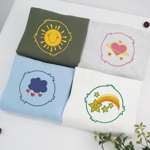 May include: Four sweatshirts in olive green, light blue, white, and gray. Each sweatshirt features a different embroidered design: a smiling sun, a heart with planets, a cloud with hearts, and a star with a rainbow.