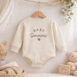 May include: A long-sleeved cream-colored baby bodysuit featuring the embroidered text BABY Simmons above a small heart icon. The garment is made of a soft, textured fabric and includes ribbed cuffs at the wrists and a snap closure at the bottom.