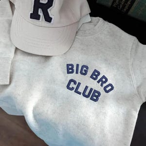 May include: A light gray sweatshirt with the text "BIG BRO CLUB" in navy blue, paired with a light gray baseball cap featuring a navy blue "R" on the front. The items are casual wear.