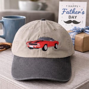 May include: A beige and grey baseball cap with an embroidered red sports car design. The cap has a curved peak and a dark grey crown. A Father's Day card and a gift box are visible in the background.
