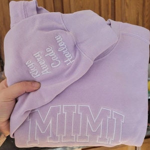 以下が含まれることがあります： 前面に大きな白いブロック文字で「MIMI」と書かれた薄紫色のパーカー。袖には「Rhys, Avery, Cade, Harlow」という名前が白で刺繍されています。パーカーは折りたたまれています。