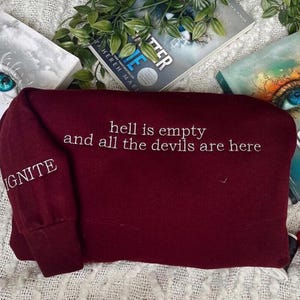 Puede incluir: Una sudadera burdeos con la frase bordada en blanco "hell is empty and all the devils are here". La palabra "IGNITE" está bordada en la manga. La sudadera está doblada sobre una superficie blanca texturizada, con libros y vegetación en el fondo.