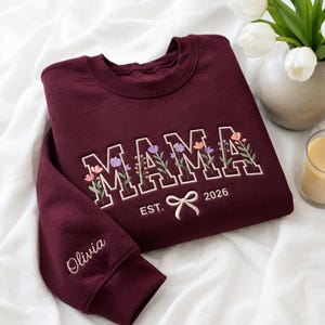 Könnte beinhalten: Ein bordeauxrotes Sweatshirt mit Rundhalsausschnitt und dem gestickten Schriftzug MAMA in weißen Blockbuchstaben, die mit bunten Blumenmotiven gefüllt sind. Unter dem Text befindet sich die Stickerei EST. 2026 mit einer kleinen Schleife. Der Name Olivia ist auf dem Ärmel eingestickt.