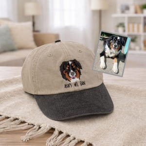 以下が含まれることがあります： ダークグレーのつばが付いたベージュの野球帽。三色の犬の刺繍ポートレートと黒い糸で「ASH'S #1 FAN」という文字が施されています。帽子の右側には、同じ犬の小さな長方形の写真が添えられています。