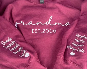 May include: A maroon long-sleeve sweatshirt featuring white embroidered text that reads "grandma EST. 2009" on the chest. The sleeves are embroidered with the names "Quade, Evander, Bennett, Briggs, Cash" and "Paislie, Henlie, Harper, Mary Kate".