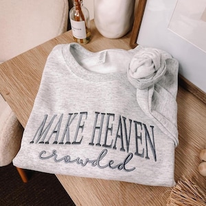 Sport Gray embroidered sweatshirt featuring the Christian phrase “Make Heaven Crowded” stitched on the chest. The words “MAKE HEAVEN” are embroidered in bold uppercase serif letters with dark gray thread, while “crowded” is stitched below in a cursive script with lighter gray thread. Minimalist unisex crewneck made from soft cotton blend, perfect for Christian apparel, faith-based outfits, church wear, or as a meaningful religious gift.