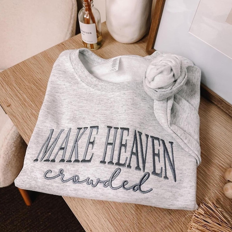 Sport Gray embroidered sweatshirt featuring the Christian phrase “Make Heaven Crowded” stitched on the chest. The words “MAKE HEAVEN” are embroidered in bold uppercase serif letters with dark gray thread, while “crowded” is stitched below in a cursive script with lighter gray thread. Minimalist unisex crewneck made from soft cotton blend, perfect for Christian apparel, faith-based outfits, church wear, or as a meaningful religious gift.