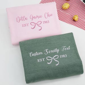 May include: Two folded sweatshirts, one pink and one green, with embroidered text and a bow design. The pink sweatshirt reads "Delta Gamie Chic EST 1983." The green sweatshirt reads "Custom Sorority Text EST 1983."