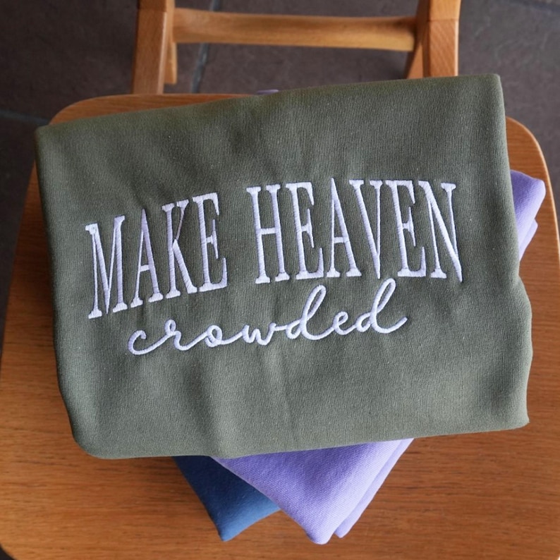 May include: An olive green sweatshirt with the embroidered phrase "MAKE HEAVEN crowded" in white script. The sweatshirt is folded and stacked on top of two other folded sweatshirts, one blue and one purple, on a wooden chair.