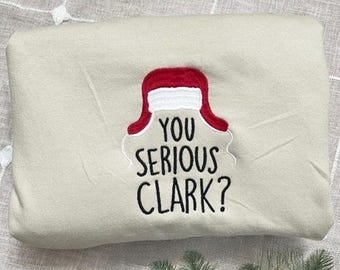 Może przedstawiać: Beżowa bluza z haftowanym wzorem przedstawiającym czerwono-białą czapkę Świętego Mikołaja i napis "YOU SERIOUS CLARK?" czarnymi literami. Złożona, ze złotymi ozdobami i zielenią na pierwszym planie.