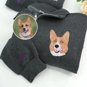 May include: Dark gray zip-up hoodie featuring a corgi dog embroidery on the chest and a smaller corgi image in a circle. The sleeve has the word "Magical" embroidered in purple. The hoodie has a silver zipper.