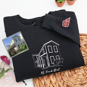 May include: Black sweatshirt featuring a white line drawing of a house with the address "112 Frank Street" printed below. A small photo of the house is on the front. A red and white logo is on the sleeve.