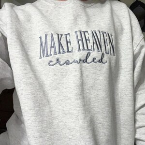 May include: A pale grey crew neck sweatshirt featuring the embroidered phrase "MAKE HEAVEN crowded" in a blue-grey hue. The text is in a stylized font. The sweatshirt is made of a soft material.