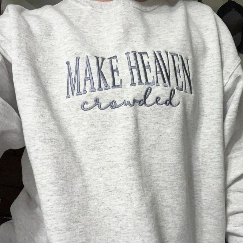 May include: A pale grey crew neck sweatshirt featuring the embroidered phrase "MAKE HEAVEN crowded" in a blue-grey hue. The text is in a stylized font. The sweatshirt is made of a soft material.