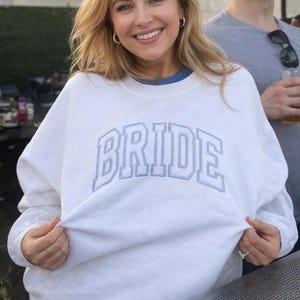 May include: White sweatshirt with the word "BRIDE" in light blue, collegiate-style lettering. The sweatshirt is being held up, showing the soft fabric. The overall theme is bridal wear.