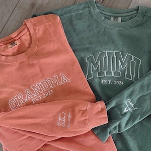 Könnte beinhalten: Zwei langärmelige Rundhals-Sweatshirts in Koralle und Salbeigrün. Das korallenfarbene Sweatshirt trägt den weißen Schriftzug "GRANDMA EST. 2023" und das salbeigrüne "MIMI EST. 2024". Beide Ärmel sind mit weiß gestickten Namen versehen.