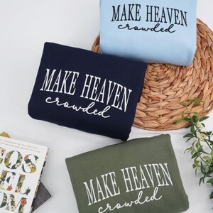 May include: Three folded sweatshirts in navy, light blue, and olive green, each with the white text "MAKE HEAVEN crowded". The sweatshirts are arranged on a woven mat with a book and greenery.