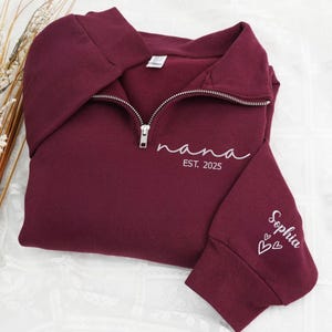 Puede incluir: Una sudadera burdeos con cremallera de un cuarto con la palabra "nana" y "EST. 2025" en escritura blanca en la parte delantera. La manga presenta el nombre "Sophia" y dos corazones blancos. La sudadera está doblada.