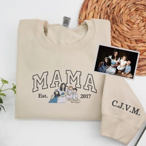 May include: A cream-coloured sweatshirt featuring the word "MAMA" in black, the text "Est. 2017", and a family illustration. A family photo is on the right. The sleeve has the initials "C.J.V.M." embroidered in black.