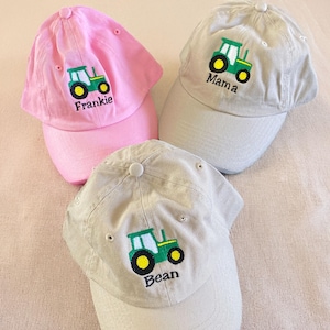 May include: Three baseball caps in pink and tan, each embroidered with a green tractor design and a name. The pink cap says "Frankie", the tan cap says "Mama", and the other tan cap says "Bean".