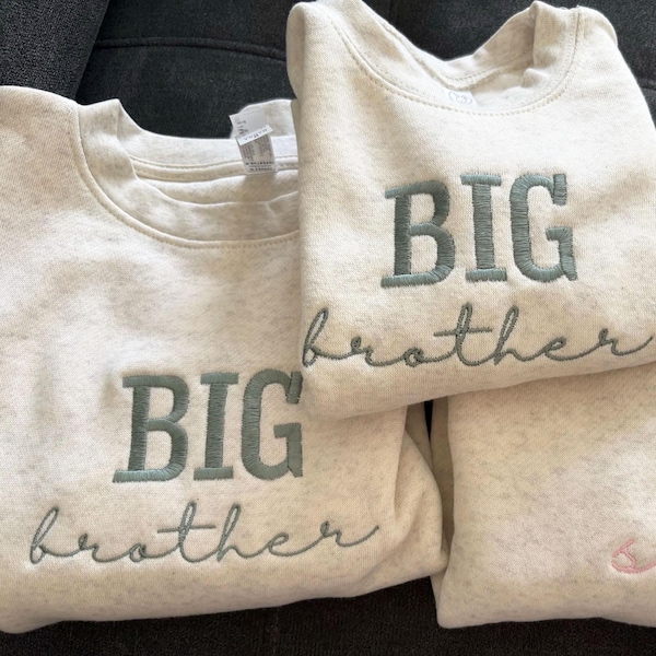 Puede incluir: Tres sudaderas de color gris claro con las palabras "BIG brother" bordadas en hilo verde azulado. Las sudaderas están dobladas y apiladas, con el texto prominentemente mostrado en la parte delantera. Una sudadera tiene un pequeño diseño bordado en rosa.