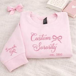Könnte beinhalten: Hellrosa Sweatshirt mit gesticktem Schriftzug "Custom Sorority" und einer Schleife. Der Ärmel zeigt den Namen "Sarah". Im Hintergrund sind eine rosa Schleife und zwei Bücher zu sehen. Der Text "est. 1895" ist ebenfalls gestickt.