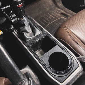 May include: Interior car detail featuring a black gear shift knob with red "TRD" lettering, a black center console with a cup holder, and a brown leather seat. The image is taken from a high angle.
