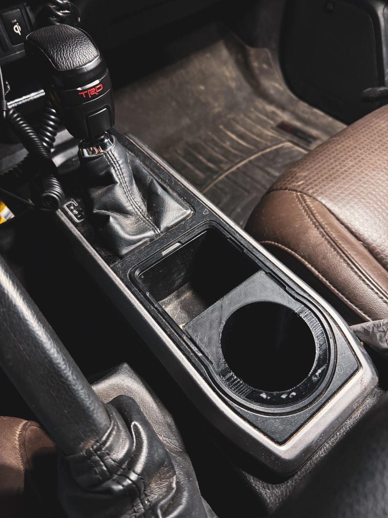 May include: Interior car detail featuring a black gear shift knob with red "TRD" lettering, a black center console with a cup holder, and a brown leather seat. The image is taken from a high angle.