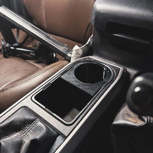 May include: Interior car console with a cup holder and storage compartment. The console is black and silver, with a leather gear shift and brown leather seats in the background. A black metal object is in the background.