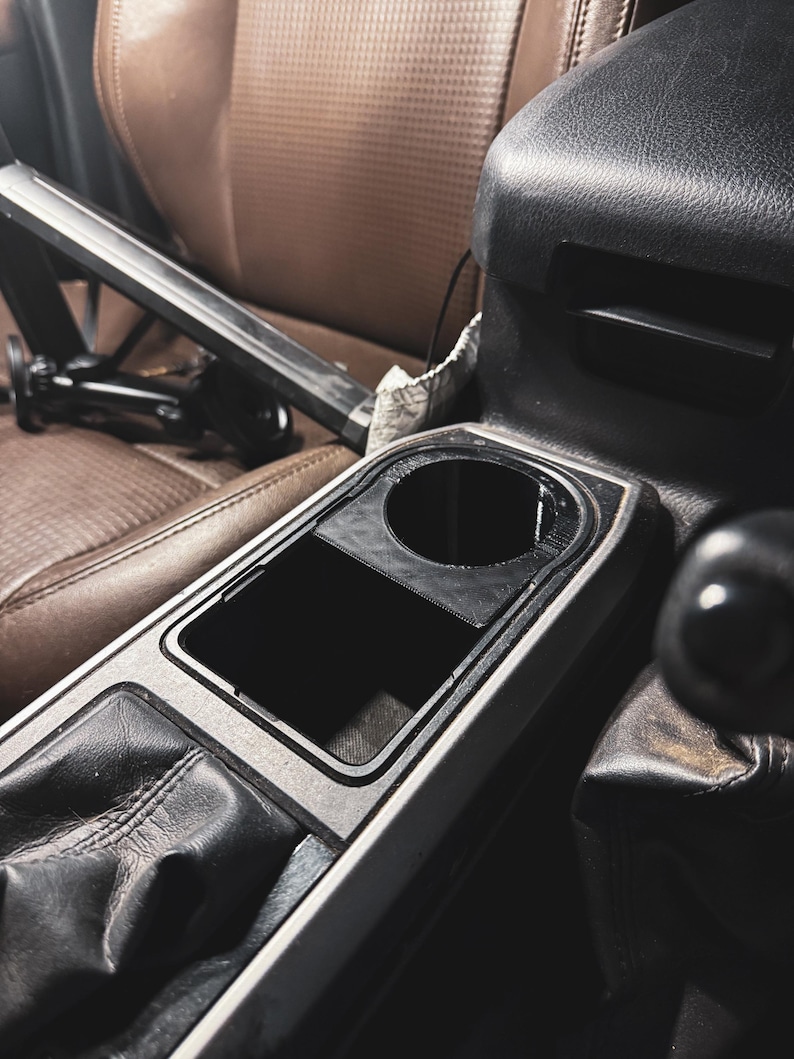 May include: Interior car console with a cup holder and storage compartment. The console is black and silver, with a leather gear shift and brown leather seats in the background. A black metal object is in the background.