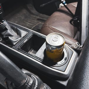 May include: A yellow and silver beverage can sits in a car's cup holder. The can has a yellow label with visible text. The interior of the car features black and brown leather and plastic components, including the gear shift and seats.
