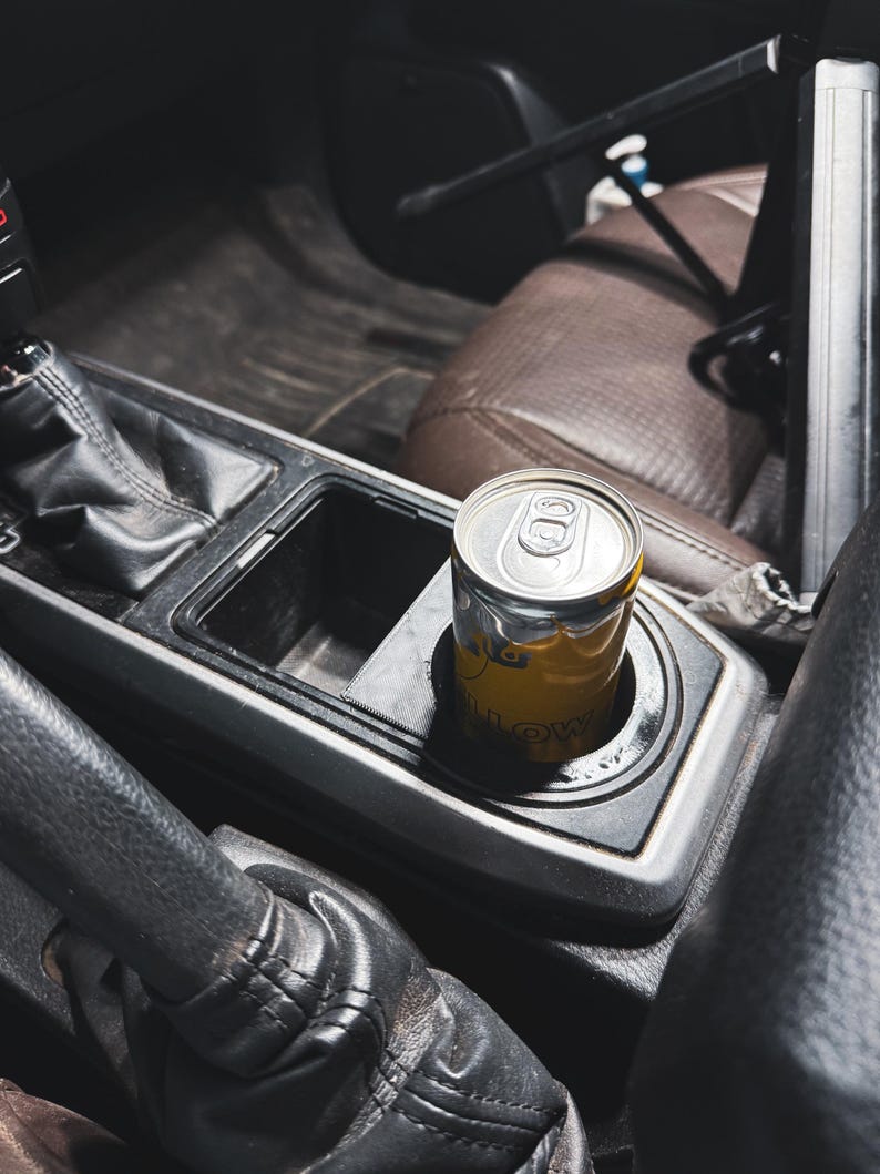 May include: A yellow and silver beverage can sits in a car's cup holder. The can has a yellow label with visible text. The interior of the car features black and brown leather and plastic components, including the gear shift and seats.
