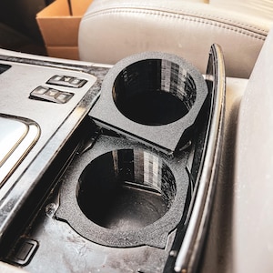 May include: Black car cup holders, designed to fit into a vehicle's centre console. The cup holders are made of a textured material and have a circular design. The image shows the cup holders installed in a car, with the car's interior visible.