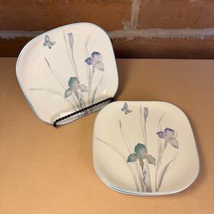 May include: Set of square, off-white plates with a light blue border. Each plate features a floral design with blue and green irises and a butterfly. The plates are displayed on a black metal plate stand.
