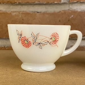 May include: A white teacup with a floral design. The cup features a rounded base, a curved handle, and a decorative pattern of red flowers and brown stems with black leaves. The cup is made of a glossy, opaque material.
