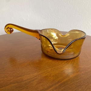 May include: Amber-colored glass serving dish with a handle. The dish has a heart-shaped bowl and a curved handle with a decorative swirl at the end. The glass is transparent, allowing light to pass through.