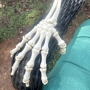 May include: A detailed, off-white skeleton hand rests on a black wicker chair. The hand is a realistic model, with individual bones and joints clearly defined. The chair is positioned outdoors, with green grass and a tree visible in the background.