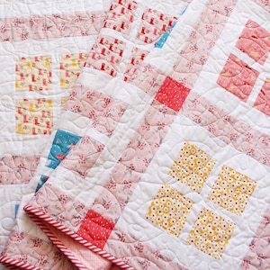 Op de afbeelding: Een roze en witte quilt met verschillende bloemenpatronen. De quilt is gemaakt van vierkanten en rechthoeken, en de randen zijn afgewerkt met een rood-wit gestreepte stof.
