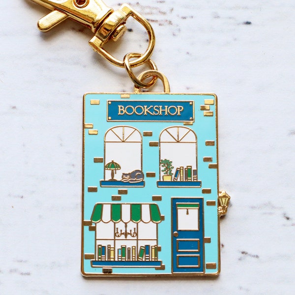 以下が含まれることがあります： 青い白の店先に「BOOKSHOP」と書かれた金色のキーホルダー。店先には窓が2つ、ドア、緑色のひさしが付いています。 
