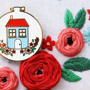 K&ouml;nnte beinhalten: Ein runder Stickrahmen aus Wei&szlig; und Gold mit einem blauen Haus mit rotem Dach und wei&szlig;en Verzierungen. Das Haus hat zwei Fenster und eine wei&szlig;e T&uuml;r. Der Rahmen ist von roten, rosa und gr&uuml;nen gestickten Blumen umgeben.