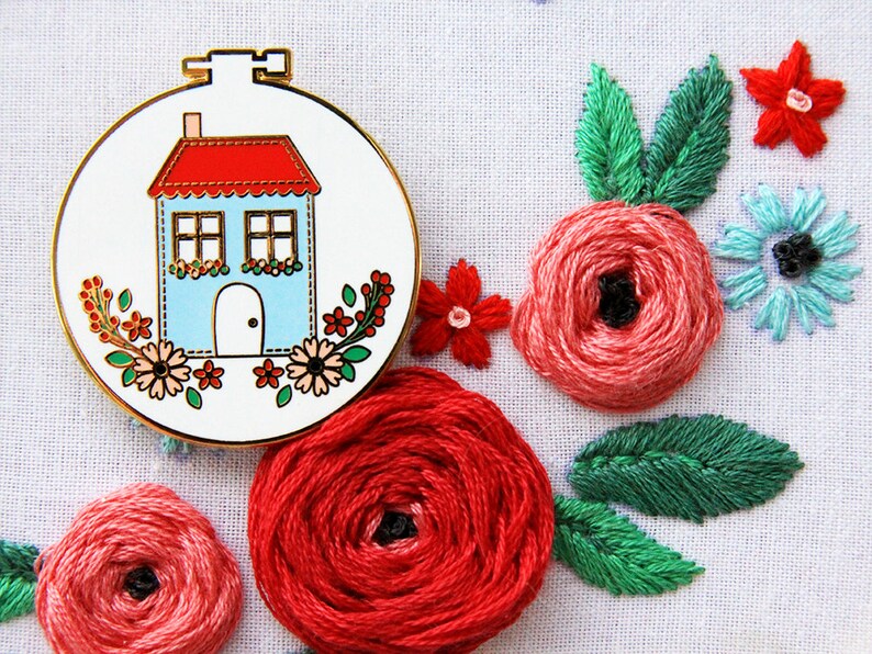K&ouml;nnte beinhalten: Ein runder Stickrahmen aus Wei&szlig; und Gold mit einem blauen Haus mit rotem Dach und wei&szlig;en Verzierungen. Das Haus hat zwei Fenster und eine wei&szlig;e T&uuml;r. Der Rahmen ist von roten, rosa und gr&uuml;nen gestickten Blumen umgeben.