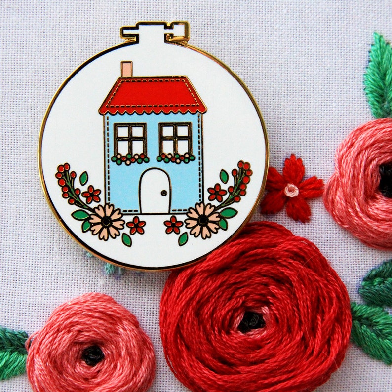 K&ouml;nnte beinhalten: Ein blaues Haus mit rotem Dach und wei&szlig;en Verzierungen ist von rosa und roten Blumen umgeben. Das Haus ist auf einen wei&szlig;en Stoffgrund gestickt.