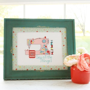 May include: A framed cross-stitch design featuring a pink sewing machine with colorful flowers and the text "Sew all the things!" The frame is teal green with a floral pattern border.