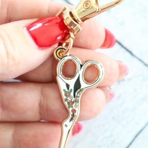 May include: Gold-tone key chain with a white enamel scissor charm decorated with colorful flowers. The scissor charm is shaped like a bird.