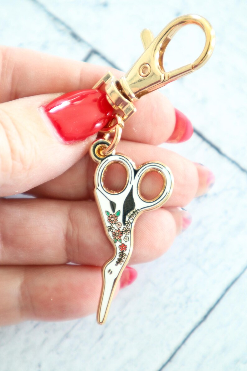 May include: Gold-tone key chain with a white enamel scissor charm decorated with colorful flowers. The scissor charm is shaped like a bird.