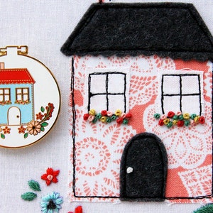 K&ouml;nnte beinhalten: Ein rosa Filzhaus mit einem grauen Filzdach, zwei Fenstern mit schwarzen Rahmen und einer schwarzen Filzt&uuml;r. Das Haus ist mit gestickten Blumen in Rosa-, Gr&uuml;n- und Gelbt&ouml;nen verziert. Das Haus steht auf einem wei&szlig;en Hintergrund.