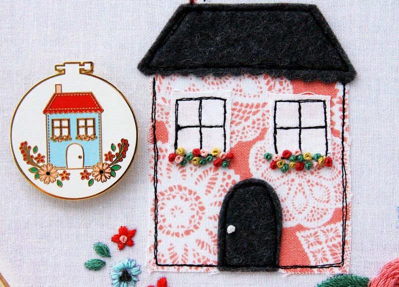 K&ouml;nnte beinhalten: Ein rosa Filzhaus mit einem grauen Filzdach, zwei Fenstern mit schwarzen Rahmen und einer schwarzen Filzt&uuml;r. Das Haus ist mit gestickten Blumen in Rosa-, Gr&uuml;n- und Gelbt&ouml;nen verziert. Das Haus steht auf einem wei&szlig;en Hintergrund.