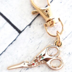 May include: Gold-tone metal key chain with a floral patterned scissor charm. The scissor charm has a white background with gold-tone metal accents and a floral pattern in shades of red, green, and brown.